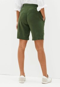 Grüne Leinenshorts mit einem Bund, Taschen auf der Rückseite und umgeschlagenen Säumen, kombiniert mit hellen Schuhen, von hinten betrachtet.