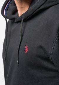 Black hoodie with adjustable drawstrings, featuring a small red logo and textured fabric. Interior shows contrasting blue and red stripes.