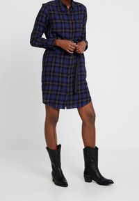Knappad rutig skjortklänning i marinblått med svarta och beige detaljer. Den har långa ärmar, bältad midja och rak skärning. Matchad med svarta stövlar.