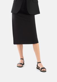 Schwarzer Bleistiftrock kombiniert mit einem schwarzen Blazer. Schwarze Sandalen mit breiten Riemen und einer Holzplattform. Glatte Texturen und klare Linien.