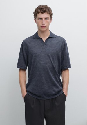 Giovane con capelli ricci che indossa una polo a maniche corte grigio scuro e pantaloni neri, in piedi con le mani in tasca davanti a uno sfondo semplice.