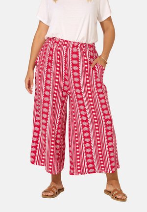 Donna che indossa pantaloni ampi a motivi rossi e bianchi, maglietta bianca a maniche corte, sandali color sabbia e braccialetti d'oro, in piedi con la mano in tasca.