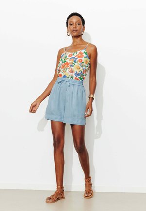 Femme portant un haut sans manches à motifs floraux, un short bleu clair, des sandales marron cloutées, des boucles d'oreilles créoles en or et un bracelet en or.