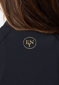Svart texturerad tygöverdel med en rund guldig logotyp som har texten "ENY" på baksidan. Har ribbat mönster och kontrasterande sömmar.