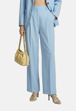Persona che indossa pantaloni azzurri chiari su misura e blazer, tiene una borsa beige con tracolla a catena dorata e indossa sandali beige con cinturini e tacchi alti.