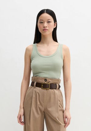 Woman standing, wearing a light green tank top tucked into high-waisted beige trousers with a dark brown belt and gold buckle.