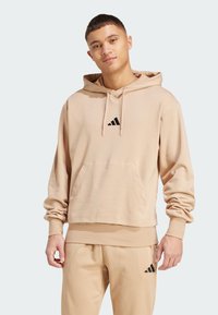 Sudadera beige con un bolsillo frontal, puños acanalados y un logo negro aplicado en el pecho. Tejido suave con un corte relajado.