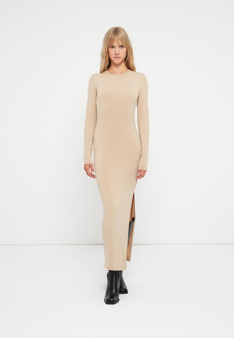 Lang beige kjole med lange ermer, sidespalte og formtilpasset design. Brukt med svarte ankelstøvler på en enkel hvit bakgrunn.