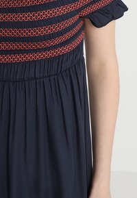 Närbild av en marinblå klänning med röda broderade geometriska mönster och korta puffärmar, synlig på en persons arm bredvid.