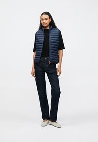 Marineblaue Steppweste mit horizontalen Steppnähten, schwarzes Hemd, dunkle Jeans und helle Schuhe. Einfaches, modernes Design mit Reißverschluss.