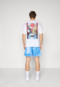 Fehér póló színes grafikus nyomatokkal, amelyek állatokat és tárgyakat ábrázolnak a hátán; kék tye-dye rövidnadrággal és fehér sportcipővel párosítva.