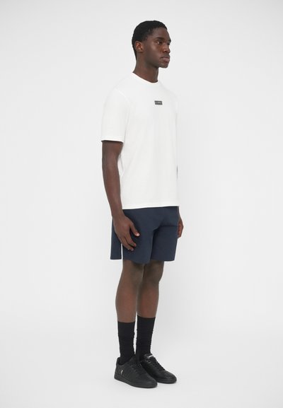 Λευκό κοντομάνικο t-shirt με ένα μικρό μαύρο λογότυπο, σε συνδυασμό με σκούρο μπλε σορτς. Το μοντέλο φοράει μαύρα αθλητικά παπούτσια και μαύρες κάλτσες μέσης.
