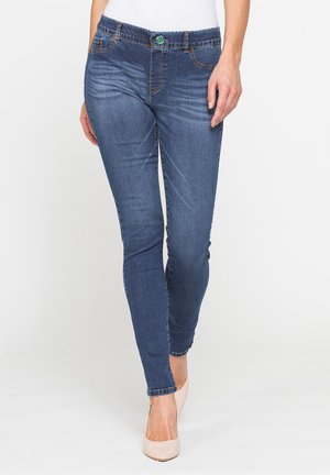 Donna che indossa jeans skinny blu e tacchi nude in piedi davanti a uno sfondo bianco con una mano appoggiata al fianco.