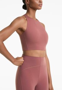 Woman wearing mauve high-neck crop top and matching high-waisted leggings, standing with one hand on her hip against white background.