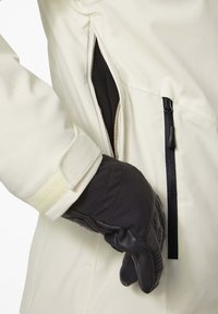 Witte soft-shell jas met een ritszak; een zwarte handschoen is te zien die in de zak reikt. De jas heeft een gestructureerde stof.