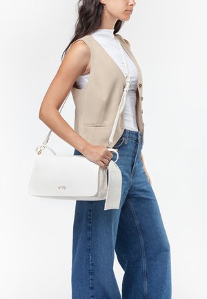 Donna che indossa un gilet beige e un top bianco tiene una borsa bianca a tracolla con tracolla decorativa, abbinata a jeans blu a gamba larga.