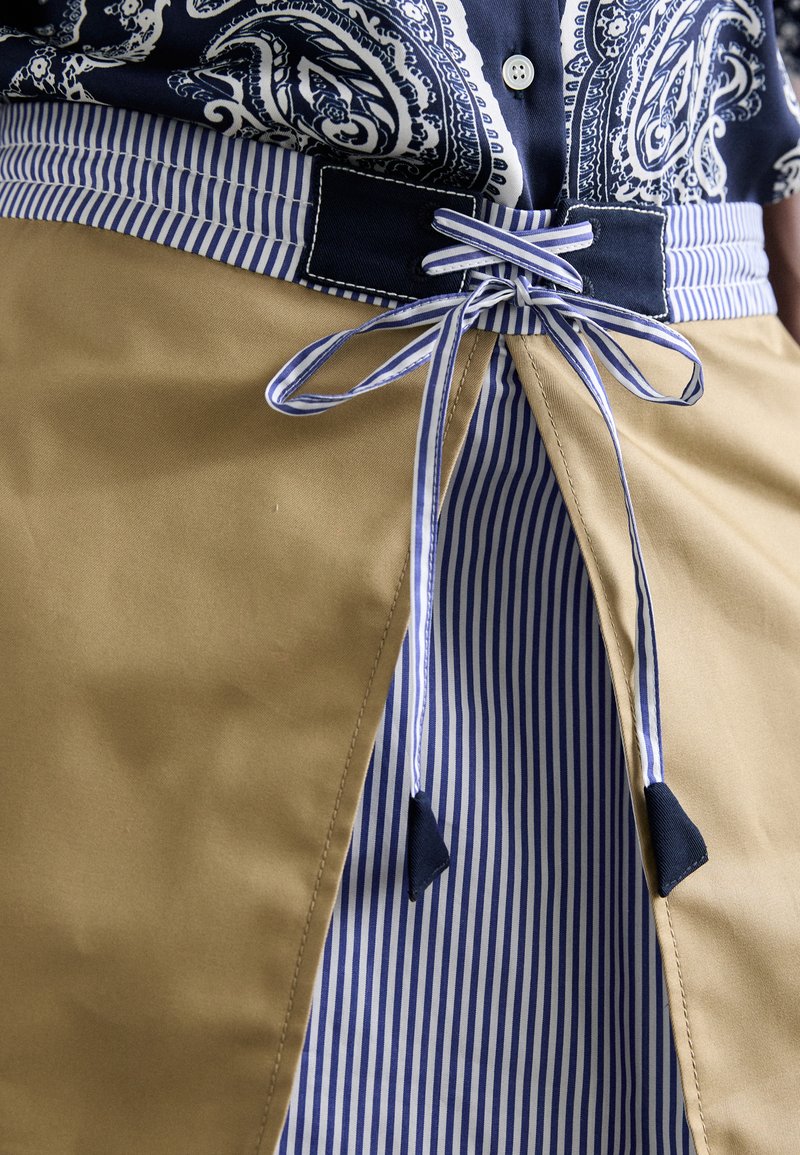 Khakifarbenes Schürze mit navy- und weißgestreiftem Taillengürtel, mit gespaltenem Vorderdesign, glatter Textur und kontrastierenden Stoffakzenten.