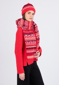 Pull en tricot rouge avec des motifs en relief en torsades, porté sous une écharpe à motifs multicolores. Bonnet rouge avec des accents de design similaires. Pantalons noirs.