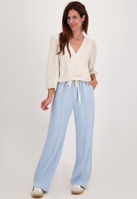 Vrouw in een crèmekleurige V-halsblouse en lichtblauwe wijde broek, staande met een hand in de zak en witte sneakers op een effen achtergrond.