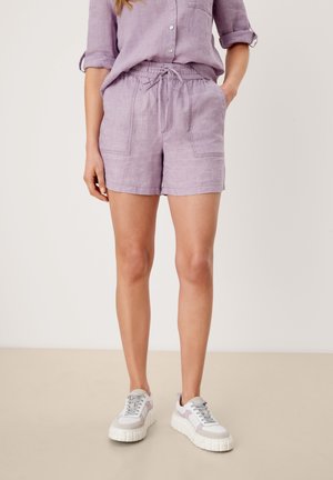 Frau trägt hellviolette Leinen-Shorts mit Fronttaschen, ein passendes Hemd zum Knöpfen mit hochgekrempelten Ärmeln und weiße Sneaker.