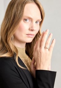 Anello in argento con pietre preziose tonde, indossato su una mano con pelle leggermente texturizzata, su uno sfondo di un blazer nero e una blusa dorata.
