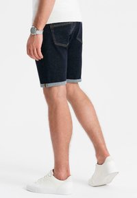 Shorts de mezclilla en azul oscuro con dobladillos enrollados, que cuentan con dos bolsillos traseros, costuras amarillas y combinados con zapatillas blancas.