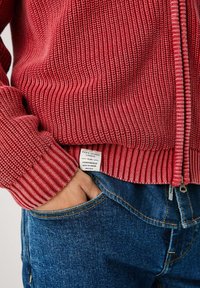 Rød ribstrikket sweater med lynlås og et stofmærke, parret med blå denim jeans, hånd i lommen, der viser det teksturerede materiale.