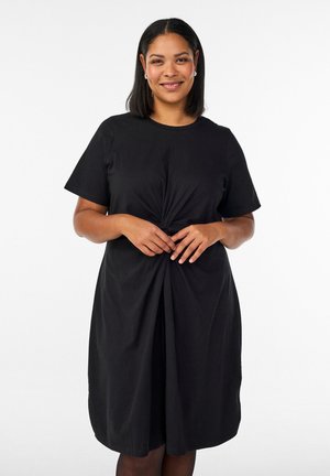 Kvinde med mellemlangt sort hår, der smiler, iført en sort kjole med korte ærmer og en knudedetalje foran, stående foran en hvid baggrund.