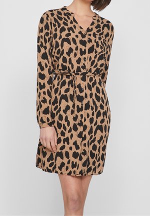 Kvinde iført en tanfarvet og sort leopardmønstret kjole med knapper, lange ærmer og et tyndt taljebånd, stående foran en ensfarvet baggrund.