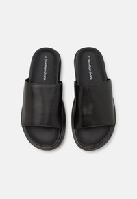 Crne klizne sandale s širokim mat trakama i mekanim đonom. Sadrže logo "Calvin Klein Jeans" na ulošku. Glatki, sintetički materijal.
