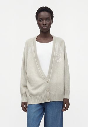 Femme aux cheveux courts et bouclés porte un cardigan beige clair boutonné sur une chemise blanche et un jean bleu, debout devant un fond uni.