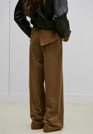 Persona con pantalones marrones de pierna ancha y chaqueta de cuero negra, de pie con las manos en las caderas sobre un fondo liso.