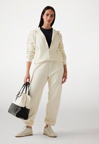 Cream zip-up hoodie with side pockets, matching cream joggers, and cream sneakers. Black-accented bag held in right hand.
