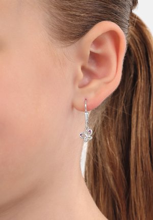 Orecchino a forma di farfalla in argento con piccoli diamanti lilla che pendono da un orecchio forato, capelli castani raccolti.