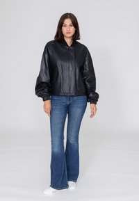 Schwarze Leder-Bomberjacke mit Reißverschluss vorne, elastischen Bündchen und seitlichen Taschen; getragen über blauen Schlagjeans und weißen Sneakern.