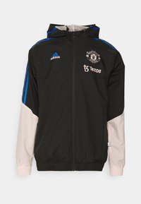 Veste de sport noire à fermeture éclair avec manches rose clair, rayures bleues sur les épaules, logo Manchester United, marquages Adidas et Tezos sur la poitrine.