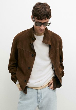 Young man wearing dark brown suede jacket, white sweater, light blue jeans, and black sunglasses, looking down with hands in pockets.