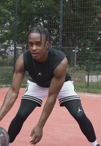 T-shirt sans manches noire, shorts blancs avec des rayures noires et leggings noirs. Ballon de basket tenu dans les mains, posture sportive sur un terrain rouge.
