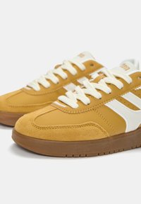 Żółte sneakersy ze zamszu i skóry z białymi akcentami, okrągłymi sznurowadłami i brązową gumową podeszwą. Zawierają detale przeszyć i logo marki.