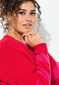 Pull rouge avec une texture côtelée et un motif de cœurs roses. Les mains du modèle sont jointes près du col, mettant en valeur le tissu doux et vibrant.