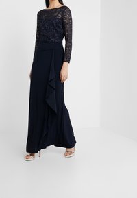 Námořnické večerní šaty s krajkovým korzetem a dlouhými rukávy, se sukní s vrstvenými volány a splývavým materiálem pro elegantní vzhled.