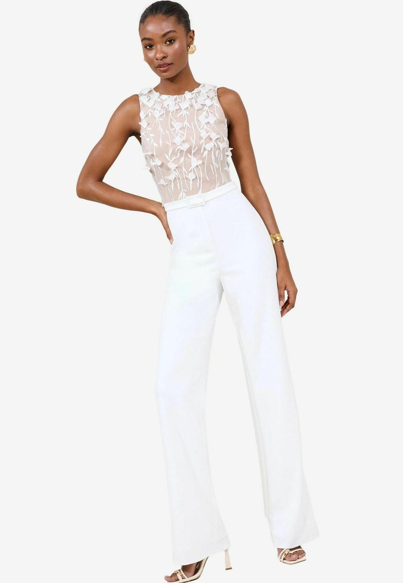 Pantalones anchos blancos combinados con una blusa sin mangas de tejido semitransparente, que presenta detalles florales bordados. El conjunto tiene un diseño moderno y elegante.