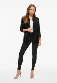 Zwarte blazer met pailletten, een sjaalkraag, driekwart mouwen en een open voorkant, gecombineerd met een wit topje en zwarte skinny broek.