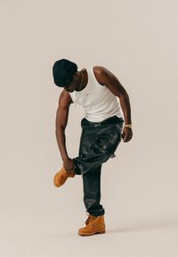 Brune semskede støvler med snørebånd, sorte skindbukser og en hvid tanktop. Personen justerer en støvle, mens de står op ad en neutral baggrund.