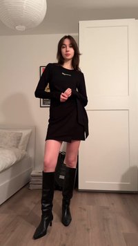 Black long-sleeve top with logo, paired with a fitted black wrap dress and knee-high black heeled boots. Wood floor and light fixture visible.