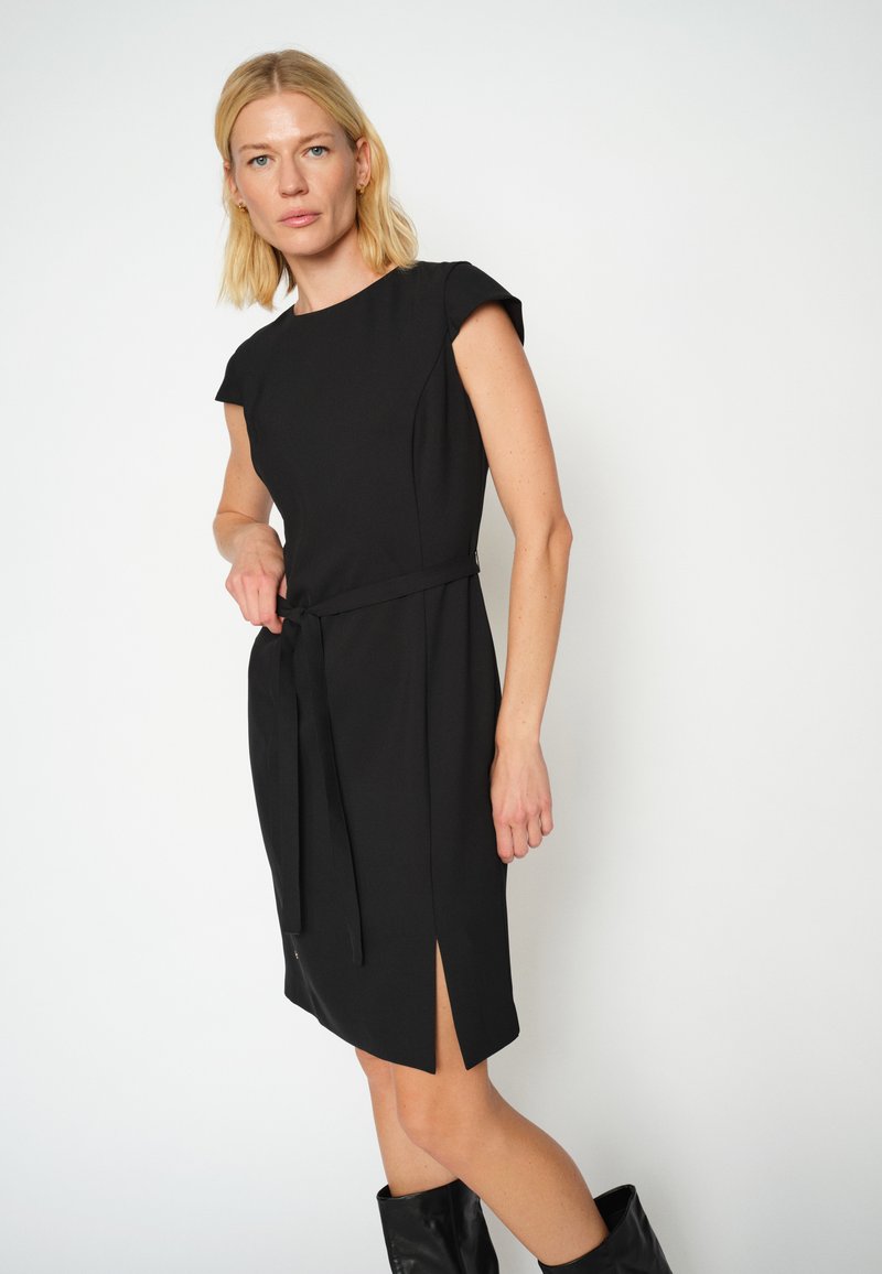 Rochie neagră cu mâneci scurte tip cap, curea în talie și fante până la coapsă. Material neted cu un design croit și un stil minimalist.