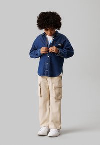 Camicia di jeans abbottonata di colore blu scuro, abbinata a pantaloni cargo beige con tasche laterali e sneakers bianche. Bambino in posizione per abbottonare la camicia.