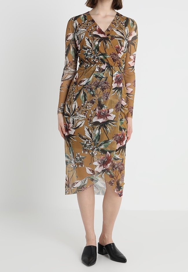 Robe portefeuille florale en marron avec manches longues. Présente un mélange de feuilles vertes et de fleurs roses, avec un ourlet mi-long. Portée avec des chaussures noires.