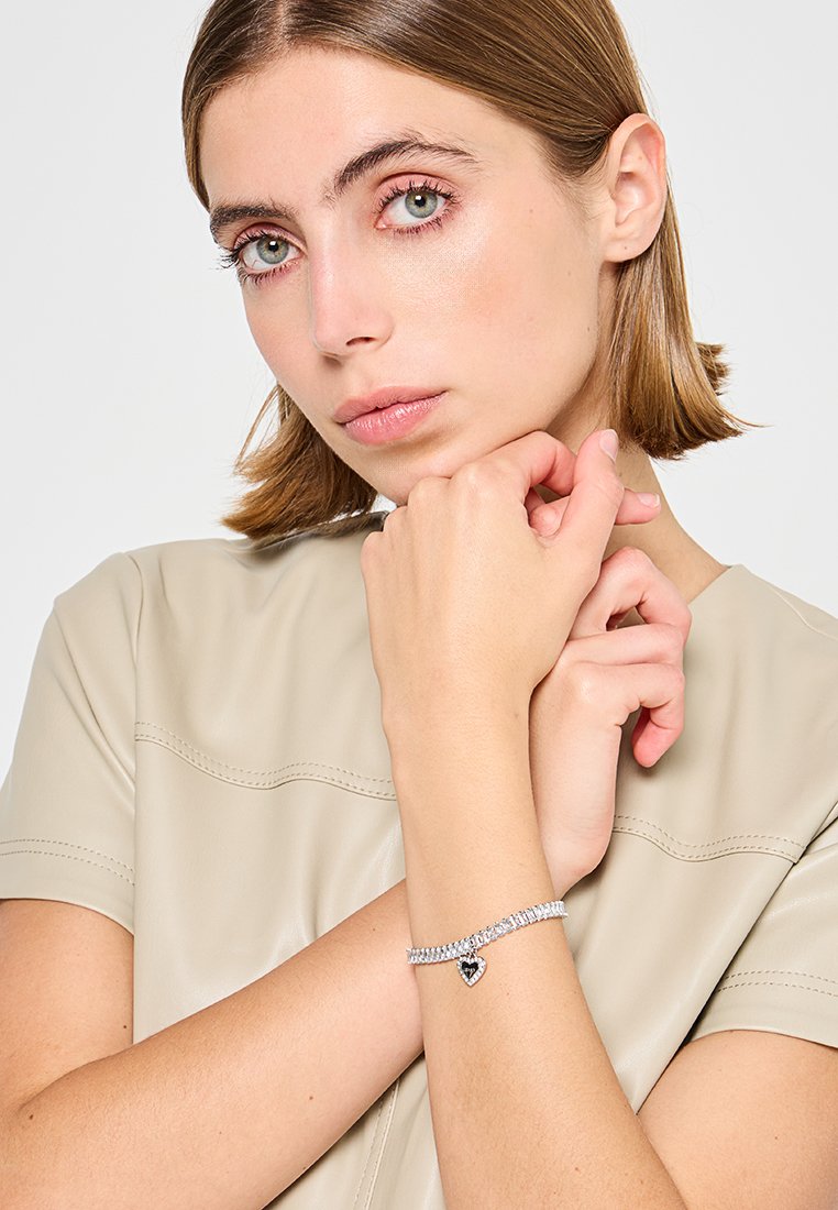 Junge Frau mit hellbraunen Haaren, die ein beiges Oberteil und ein silbernes Armband mit einem herzförmigen Anhänger am Handgelenk trägt.