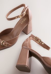 Coppia di sandali con tacco a blocco lucido color nude, con cinturini alla caviglia e fibbie dorate su sfondo chiaro.